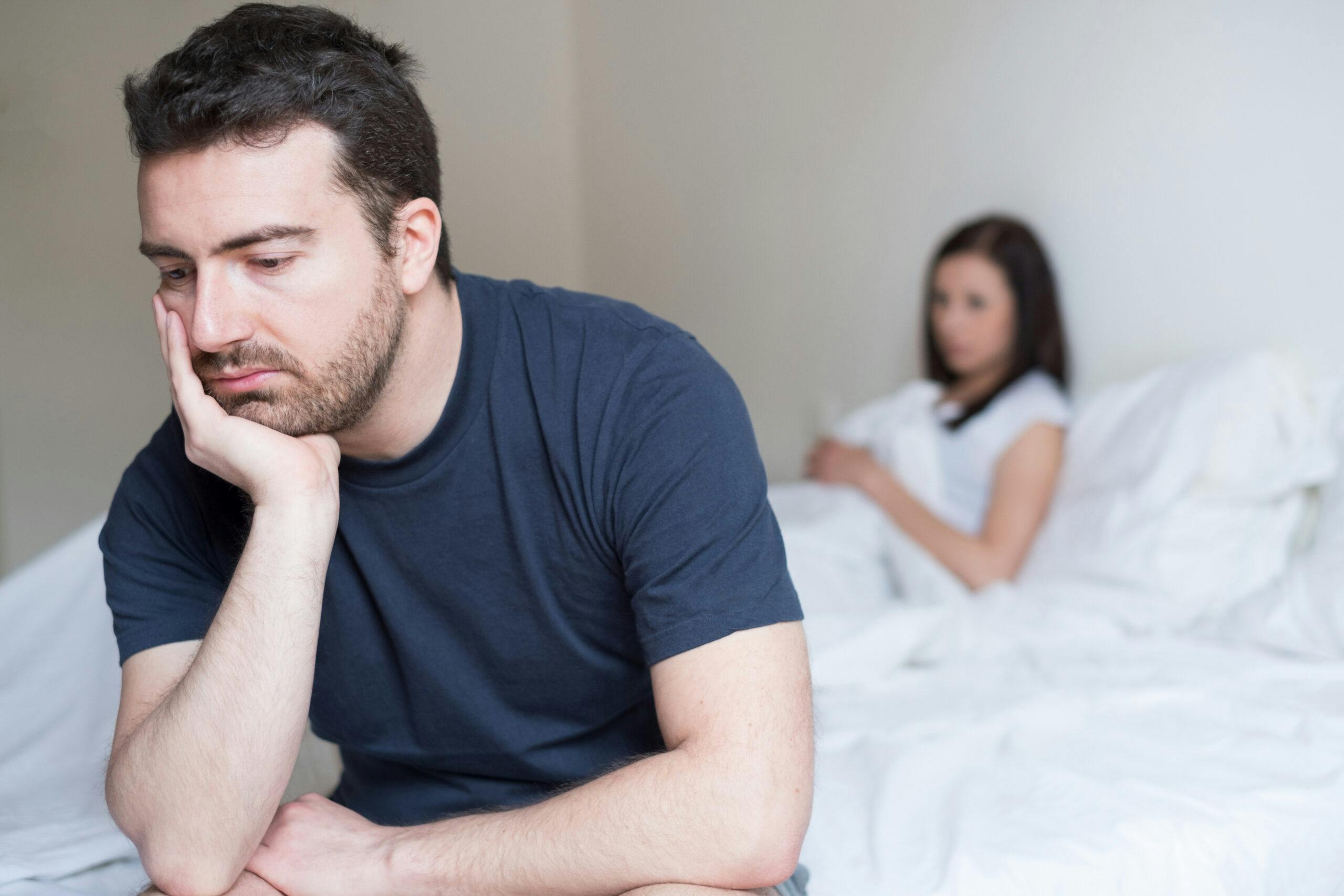 man sick upset sitting on bed with girlfriend in background
